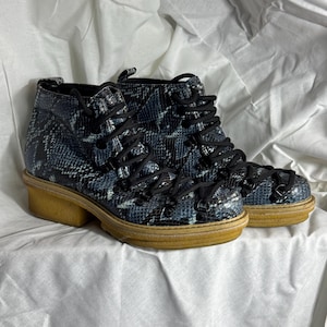 Puede incluir: Un par de botines negros y azules con estampado de piel de serpiente, con cordones negros y una suela gruesa de color marrón claro.