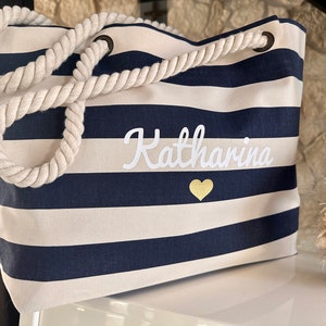 May include: A striped navy blue and white canvas tote bag with a rope handle. The bag has the name "Katharina" embroidered in white with a gold heart.