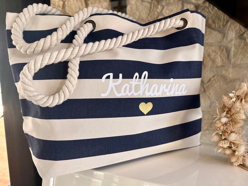 May include: A striped navy blue and white canvas tote bag with a rope handle. The bag has the name "Katharina" embroidered in white with a gold heart.