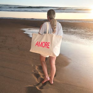 Könnte beinhalten: Eine beige Tragetasche mit dem Wort "AMORE" in roten Buchstaben. Die Tasche hat cremefarbene Träger und wird an einem Strand getragen. Die Person trägt ein gestreiftes Hemd und Shorts.