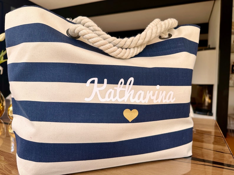 May include: A white and navy blue striped tote bag with a rope handle. The bag has the name "Katharina" embroidered in white with a gold heart below it.