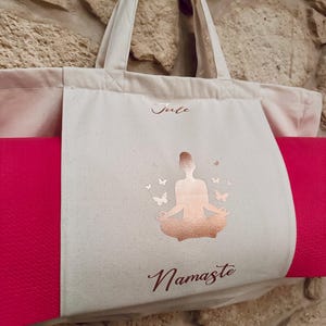 Puede incluir: Una bolsa de lona blanca con una esterilla de yoga rosa en su interior. La bolsa tiene un dise&ntilde;o en oro rosa de una mujer en una postura de yoga con mariposas y la palabra "Namaste". La bolsa tambi&eacute;n tiene el nombre "Julie" escrito en ella.