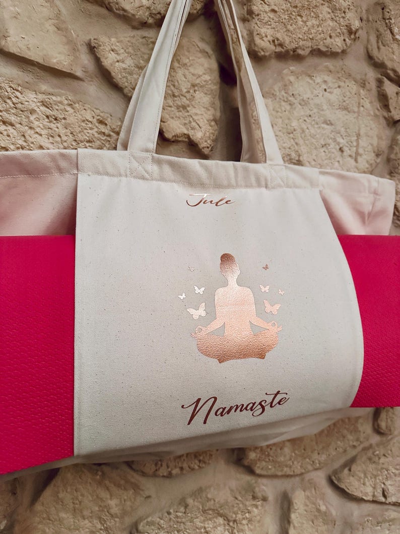 Puede incluir: Una bolsa de lona blanca con una esterilla de yoga rosa en su interior. La bolsa tiene un dise&ntilde;o en oro rosa de una mujer en una postura de yoga con mariposas y la palabra "Namaste". La bolsa tambi&eacute;n tiene el nombre "Julie" escrito en ella.