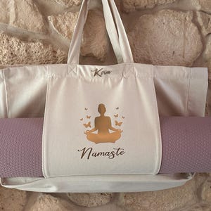 Puede incluir: Una bolsa de lona blanca con una esterilla de yoga morada en su interior. La bolsa de lona tiene un bolsillo en la parte delantera con una silueta dorada de una persona en una postura de yoga y la palabra "Namaste" impresa debajo. La bolsa de lona tiene el nombre "Kira" escrito en la parte superior.