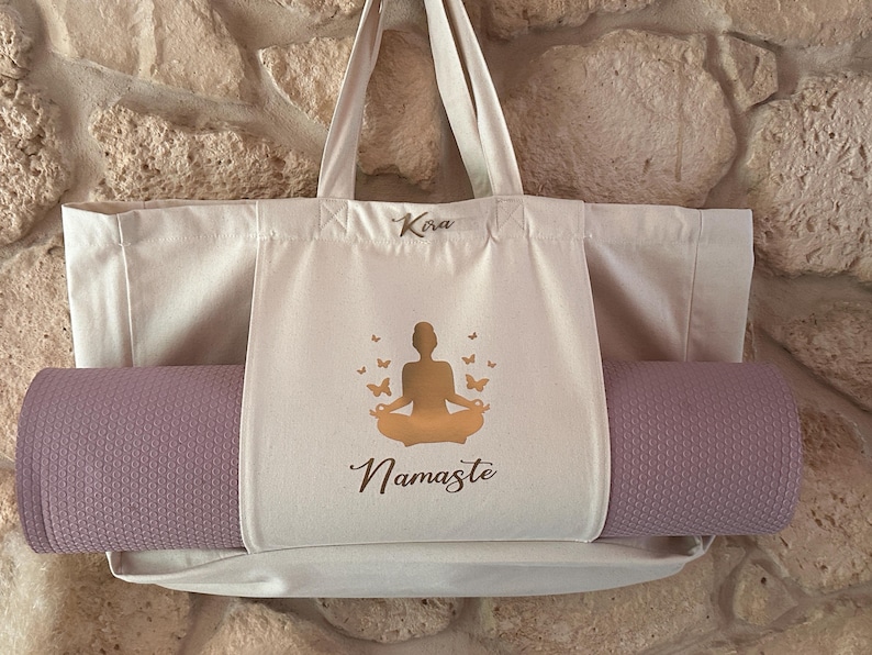 Puede incluir: Una bolsa de lona blanca con una esterilla de yoga morada en su interior. La bolsa de lona tiene un bolsillo en la parte delantera con una silueta dorada de una persona en una postura de yoga y la palabra "Namaste" impresa debajo. La bolsa de lona tiene el nombre "Kira" escrito en la parte superior.