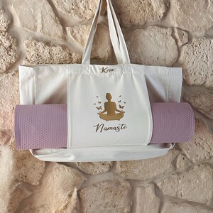 Puede incluir: Una bolsa de lona blanca con una esterilla de yoga en su interior. La bolsa tiene una silueta dorada de una persona en una postura de yoga con mariposas y la palabra "Namaste" impresa en ella. La bolsa tambi&eacute;n tiene la palabra "Kira" impresa en ella.