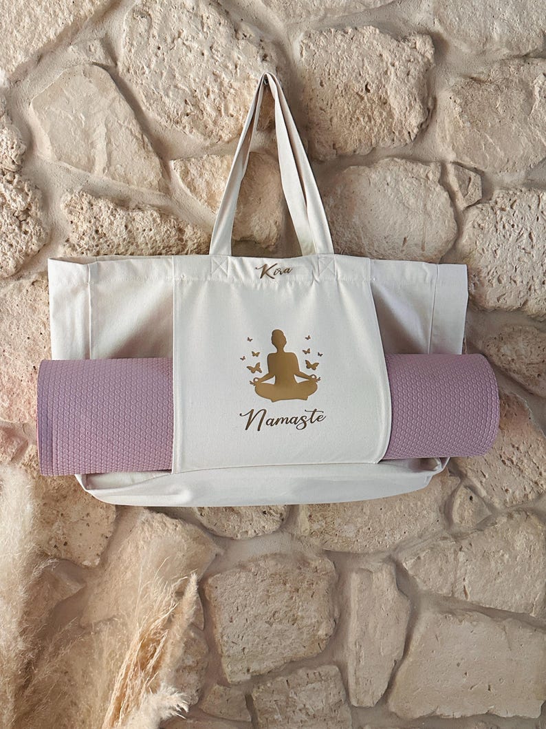 Puede incluir: Una bolsa de lona blanca con una esterilla de yoga en su interior. La bolsa tiene una silueta dorada de una persona en una postura de yoga con mariposas y la palabra "Namaste" impresa en ella. La bolsa tambi&eacute;n tiene la palabra "Kira" impresa en ella.