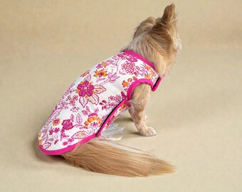 Puede incluir: Un perro pequeño con un chaleco para perros con estampado floral. El chaleco presenta un fondo blanco con flores rosas y naranjas y un ribete rosa brillante. El perro está de espaldas a la cámara.