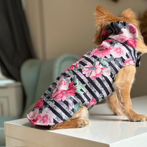 K&ouml;nnte beinhalten: Ein kleiner Hund tr&auml;gt eine schwarz-wei&szlig; gestreifte Kapuzenjacke mit Blumenmuster. Die Jacke hat rosa und wei&szlig;e Blumen auf schwarzem Hintergrund.