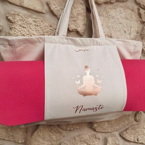 Puede incluir: Una bolsa de lona blanca con una esterilla de yoga rosa en su interior. La bolsa tiene un dise&ntilde;o de l&aacute;mina dorada de una mujer en una postura de yoga con mariposas y el texto "Namaste".