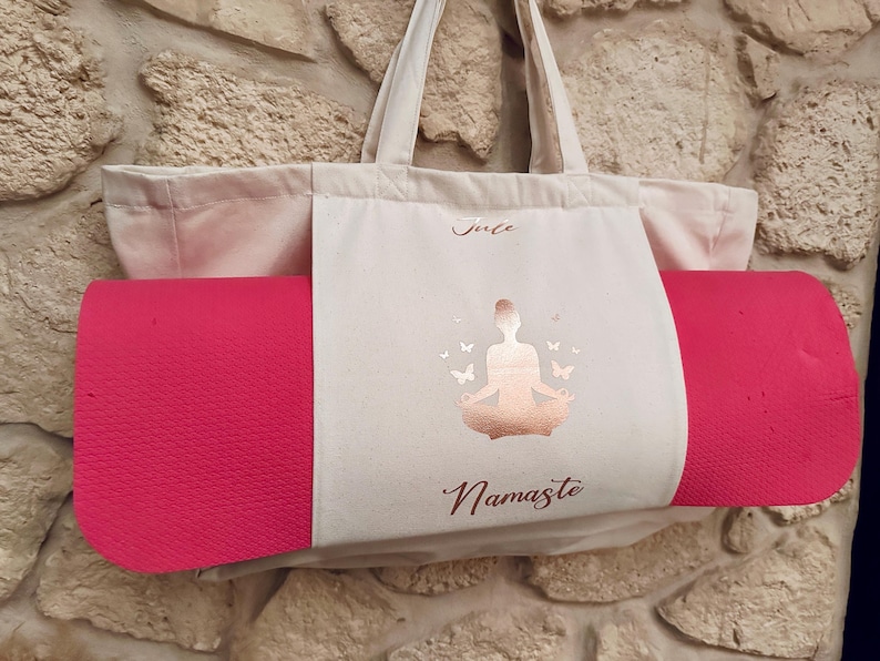 Puede incluir: Una bolsa de lona blanca con una esterilla de yoga rosa en su interior. La bolsa tiene un dise&ntilde;o de l&aacute;mina dorada de una mujer en una postura de yoga con mariposas y el texto "Namaste".