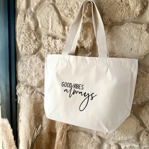 Könnte beinhalten: Eine weiße Canvas-Tragetasche mit schwarzem Text, der "GOOD VIBES always" lautet. Die Tasche hängt an einer Steinwand.