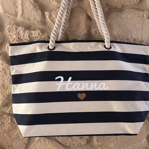 May include: A white and navy blue striped tote bag with rope handles. The bag has the name "Hanna" written in white cursive with a gold heart below it.