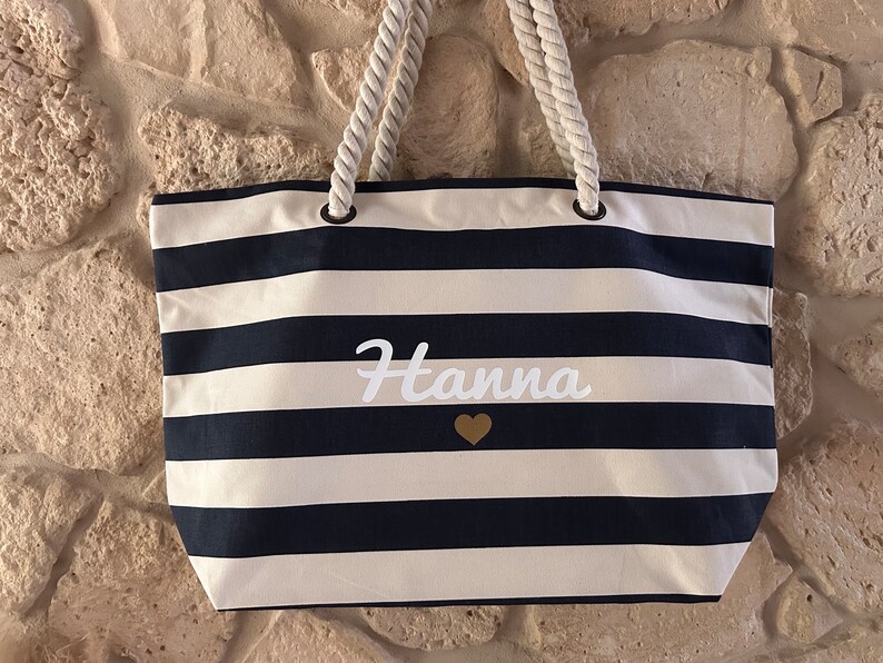 May include: A white and navy blue striped tote bag with rope handles. The bag has the name "Hanna" written in white cursive with a gold heart below it.