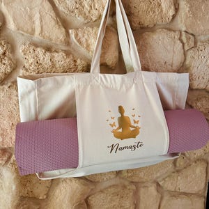 Puede incluir: Una bolsa de lona beige con una esterilla de yoga metida dentro. La bolsa tiene una silueta dorada de una persona en una postura de yoga y la palabra "Namaste" impresa en ella.