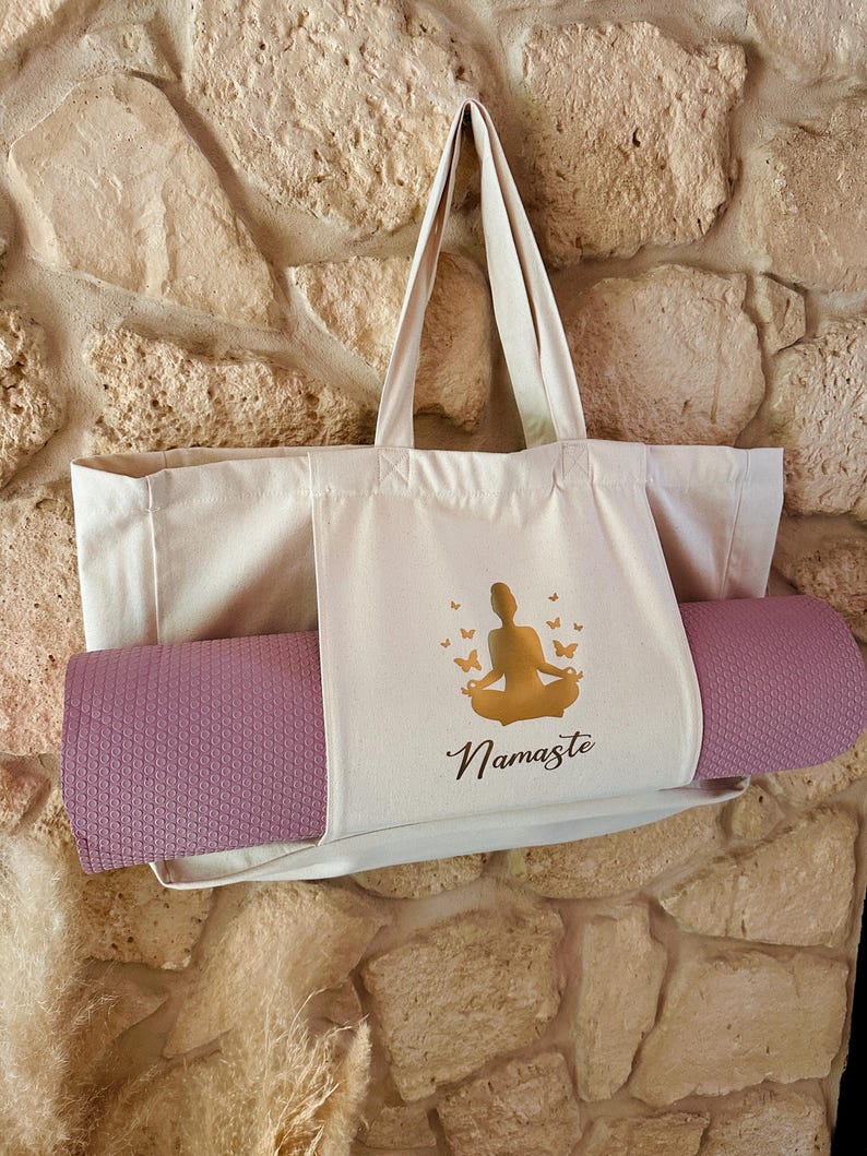 Puede incluir: Una bolsa de lona beige con una esterilla de yoga metida dentro. La bolsa tiene una silueta dorada de una persona en una postura de yoga y la palabra "Namaste" impresa en ella.