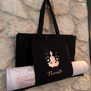 Puede incluir: Bolsa de yoga negra con una esterilla de yoga rosa y blanca en su interior. La bolsa tiene una silueta de color rosa dorado de una persona meditando con mariposas y la palabra "Namaste" impresa en ella.