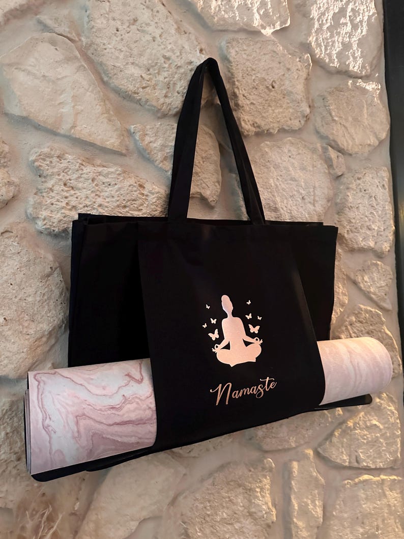 Puede incluir: Bolsa de yoga negra con una esterilla de yoga rosa y blanca en su interior. La bolsa tiene una silueta de color rosa dorado de una persona meditando con mariposas y la palabra "Namaste" impresa en ella.