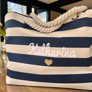 May include: A white and navy blue striped tote bag with a rope handle. The bag has the name "Katharina" embroidered on it in white with a gold heart below the name.