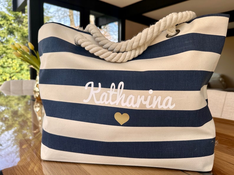 May include: A white and navy blue striped tote bag with a rope handle. The bag has the name "Katharina" embroidered on it in white with a gold heart below the name.