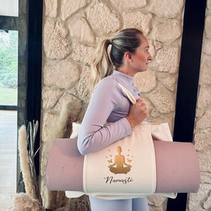 Puede incluir: Una bolsa blanca para tapete de yoga con una silueta de yoga dorada y mariposas. La bolsa tiene la palabra "Namaste" impresa. Una persona sostiene la bolsa con un tapete de yoga dentro.