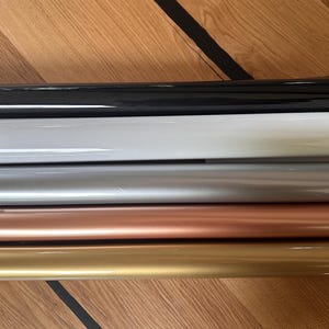 Puede incluir: Cinco rollos de pel&iacute;cula adhesiva de vinilo en diferentes colores: negro, blanco, plateado, oro rosa y oro. Los rollos est&aacute;n apilados uno encima del otro sobre una superficie de madera.