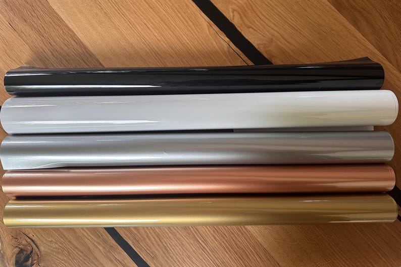 Puede incluir: Cinco rollos de pel&iacute;cula adhesiva de vinilo en diferentes colores: negro, blanco, plateado, oro rosa y oro. Los rollos est&aacute;n apilados uno encima del otro sobre una superficie de madera.