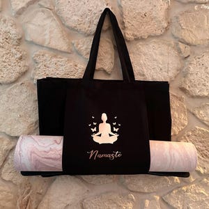 Puede incluir: Bolsa de yoga negra con una silueta blanca de una persona meditando con mariposas a su alrededor. El texto "Namaste" est&aacute; impreso debajo de la silueta. Una esterilla de yoga rosa y blanca est&aacute; metida en la bolsa.