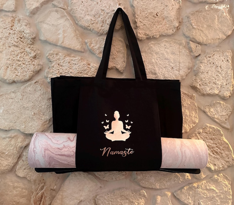 Puede incluir: Bolsa de yoga negra con una silueta blanca de una persona meditando con mariposas a su alrededor. El texto "Namaste" est&aacute; impreso debajo de la silueta. Una esterilla de yoga rosa y blanca est&aacute; metida en la bolsa.