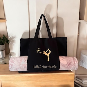 Könnte beinhalten: Schwarze Yogatasche mit einer goldenen Yoga-Silhouette und Schmetterlingen. Die Tasche enthält eine rosa-weiße marmorierte Yogamatte. Der Text auf der Tasche lautet "Katha's Yoga vibes only".