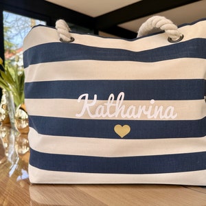 May include: A white and navy blue striped tote bag with a rope handle. The bag has the name "Katharina" embroidered in white with a gold heart.
