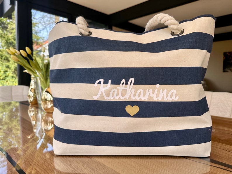 May include: A white and navy blue striped tote bag with a rope handle. The bag has the name "Katharina" embroidered in white with a gold heart.