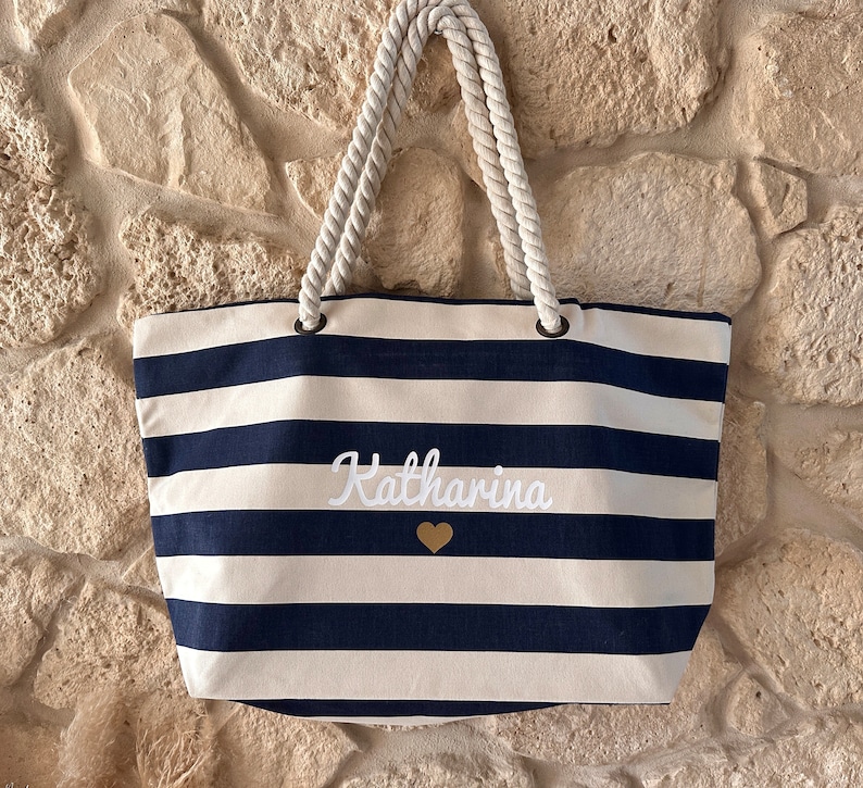 May include: A white and navy blue striped tote bag with rope handles. The bag has the name "Katharina" embroidered on the front in white with a gold heart.
