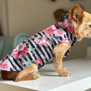 K&ouml;nnte beinhalten: Ein kleiner, hellbrauner Hund, der einen schwarz-wei&szlig; gestreiften Kapuzenregenmantel mit rosafarbenem Blumenmuster tr&auml;gt. Der Regenmantel hat eine Kapuze und ist f&uuml;r kleine Hunde konzipiert.