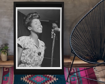Puede incluir: Una fotografía enmarcada en blanco y negro que muestra a una persona con el pelo corto sonriendo mientras canta ante un micrófono con la etiqueta WMCA. La persona viste una blusa de manga corta con un estampado floral y un broche circular.