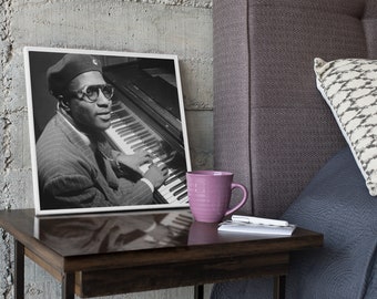 Puede incluir: Una fotografía enmarcada en blanco y negro de un músico con boina y gafas tocando el piano. El marco cuadrado es blanco y se apoya sobre una superficie de madera oscura junto a una taza de cerámica morada y un bolígrafo blanco sobre una libreta.