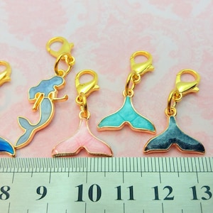 Peut inclure: Cinq breloques de queue de sirène dorées avec des détails en émail. Les breloques sont bleues, roses, turquoise et deux nuances de bleu. Les breloques sont suspendues à des fermoirs à ressort dorés. Une règle est en arrière-plan.