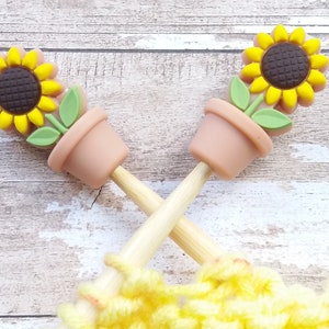 Peut inclure: Deux aiguilles à tricoter en bois avec des décorations en forme de pot de tournesol. Les aiguilles sont croisées et ont du fil jaune enroulé autour d'elles.