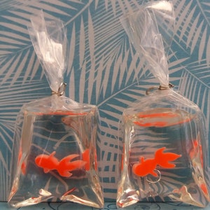 K&ouml;nnte beinhalten: Zwei transparente Plastikt&uuml;ten, jede mit einem orangefarbenen Goldfisch-Anh&auml;nger in klarer Fl&uuml;ssigkeit. Die Beutel sind mit einem silberfarbenen Metallring verschlossen und mit klarem Kunststoff gebunden. Der Hintergrund zeigt ein blau-wei&szlig;es tropisches Bl&auml;ttermuster.