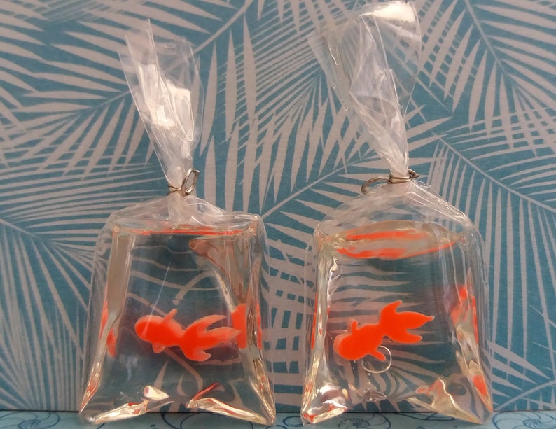 K&ouml;nnte beinhalten: Zwei transparente Plastikt&uuml;ten, jede mit einem orangefarbenen Goldfisch-Anh&auml;nger in klarer Fl&uuml;ssigkeit. Die Beutel sind mit einem silberfarbenen Metallring verschlossen und mit klarem Kunststoff gebunden. Der Hintergrund zeigt ein blau-wei&szlig;es tropisches Bl&auml;ttermuster.