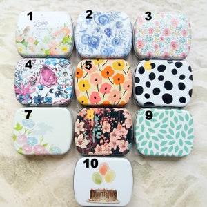 K&ouml;nnte beinhalten: Eine Sammlung von zehn kleinen, rechteckigen Metallboxen mit verschiedenen dekorativen Designs. Die Dosen zeigen florale Muster, Polka Dots und Illustrationen von Kuchen und Luftballons. Die Farbpalette umfasst Pastellt&ouml;ne, leuchtende Farben sowie Schwarz und Wei&szlig;.