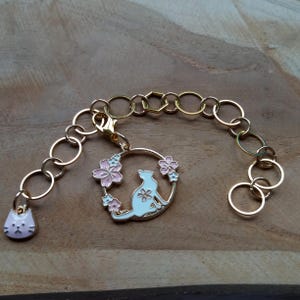 Könnte beinhalten: Ein goldfarbenes Armband mit einem Katzenanhänger. Der Katzenanhänger ist von rosa und blauen Blumen umgeben. Das Armband besteht aus einer Kette mit einem Karabinerverschluss.