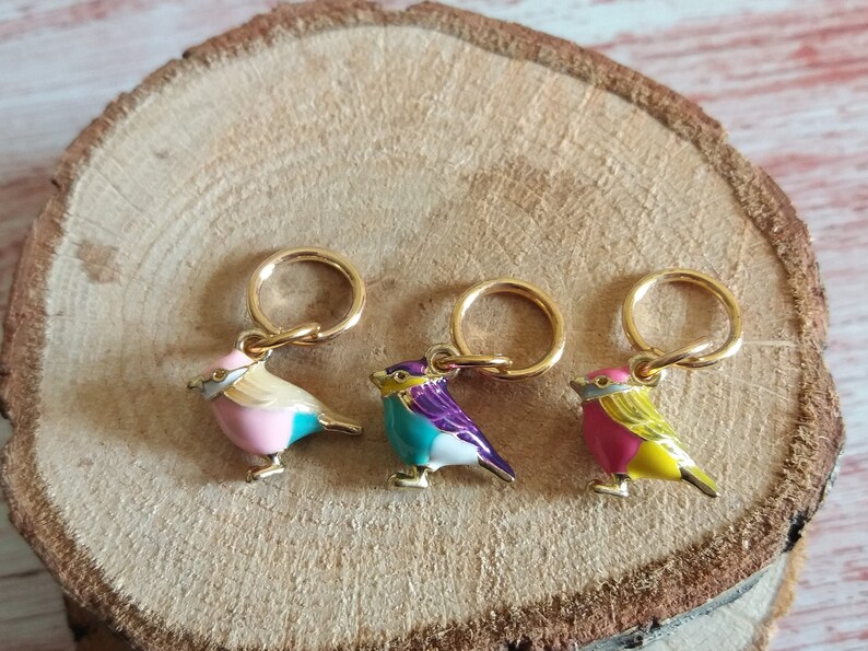 May include: Three gold stitch markers with colorful bird charms. The birds are pink and teal, teal and purple, and pink and yellow.