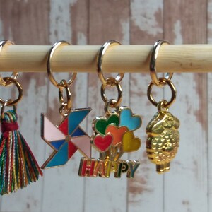 Pu&ograve; includere: Cinque marcapunti colorati con diversi design. I marcapunti sono attaccati a un bastoncino di legno. I design includono un fiocco, una girandola, un mazzo di palloncini con la scritta "HAPPY" sotto e un uccello dorato.