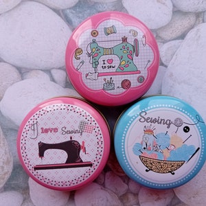 Op de afbeelding: Drie ronde, decoratieve naaidozen. E&eacute;n roze doos heeft een naaimachine-afbeelding met de tekst "I love to sew". Een andere roze doos heeft een naaimachine-afbeelding met de tekst "I love Sewing". Een blauwe doos heeft een naaithema.
