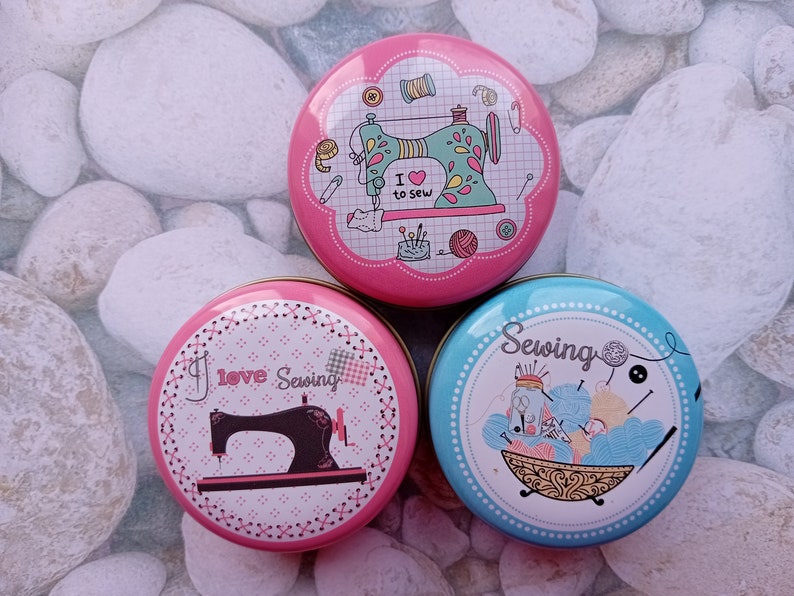 Op de afbeelding: Drie ronde, decoratieve naaidozen. E&eacute;n roze doos heeft een naaimachine-afbeelding met de tekst "I love to sew". Een andere roze doos heeft een naaimachine-afbeelding met de tekst "I love Sewing". Een blauwe doos heeft een naaithema.
