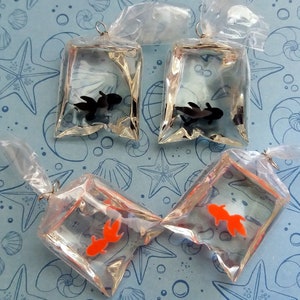 K&ouml;nnte beinhalten: Vier transparente Plastikt&uuml;ten, jede mit einem kleinen, dekorativen Fisch. Zwei T&uuml;ten enthalten schwarze Fische, zwei orangefarbene Fische. Die T&uuml;ten sind oben mit einem silbernen Draht gebunden, auf blauem Hintergrund mit Muschelmuster.