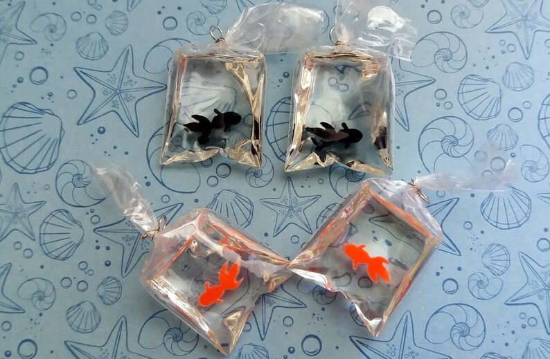 K&ouml;nnte beinhalten: Vier transparente Plastikt&uuml;ten, jede mit einem kleinen, dekorativen Fisch. Zwei T&uuml;ten enthalten schwarze Fische, zwei orangefarbene Fische. Die T&uuml;ten sind oben mit einem silbernen Draht gebunden, auf blauem Hintergrund mit Muschelmuster.