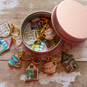 May include: A set of colorful tea-themed stitch markers in a pink metal tin. The markers feature teacups, teapots, and houses with the text "A Cup of Tea Solves Everything".