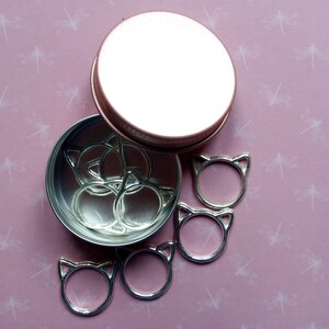 May include: A set of silver cat-shaped stitch markers in a round metal tin with a rose gold lid. The markers are arranged in the tin and some are scattered around it.